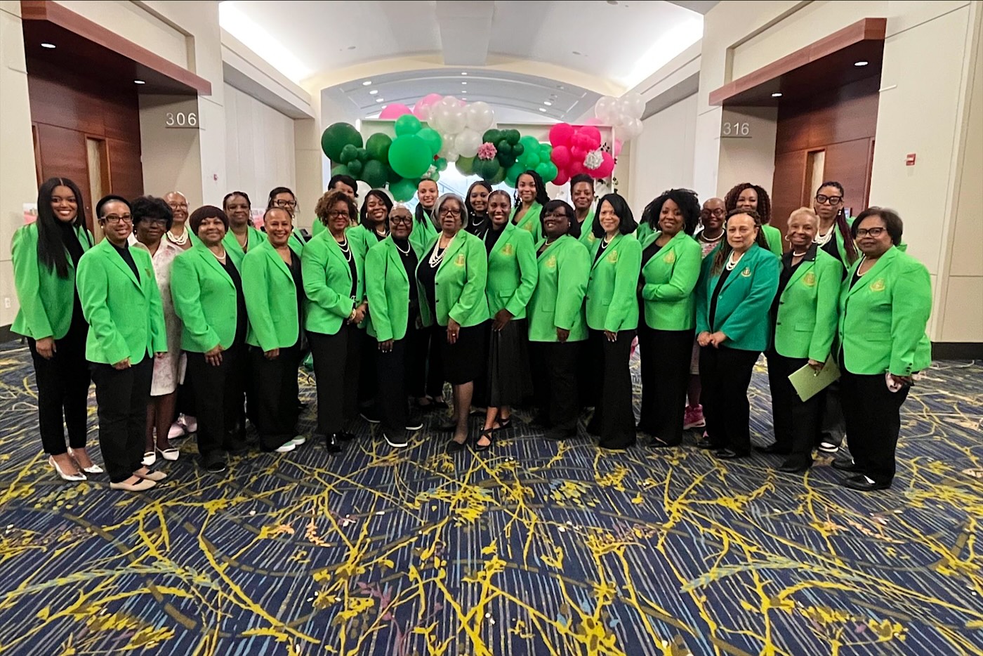 Iota Zeta Omega sorors in green blazers in front of pink, green, and white balloons